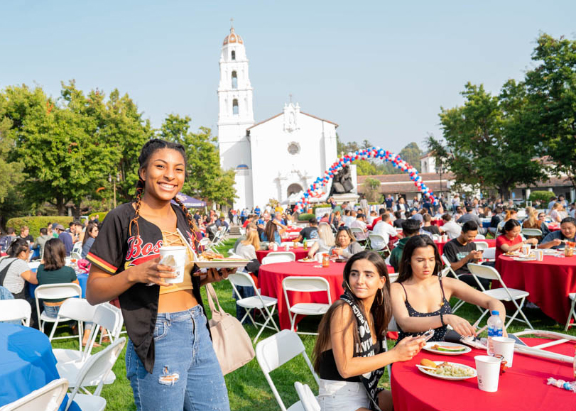 A girl standing up with a plate and cup in her hand smiling during an event on the chapel lawn