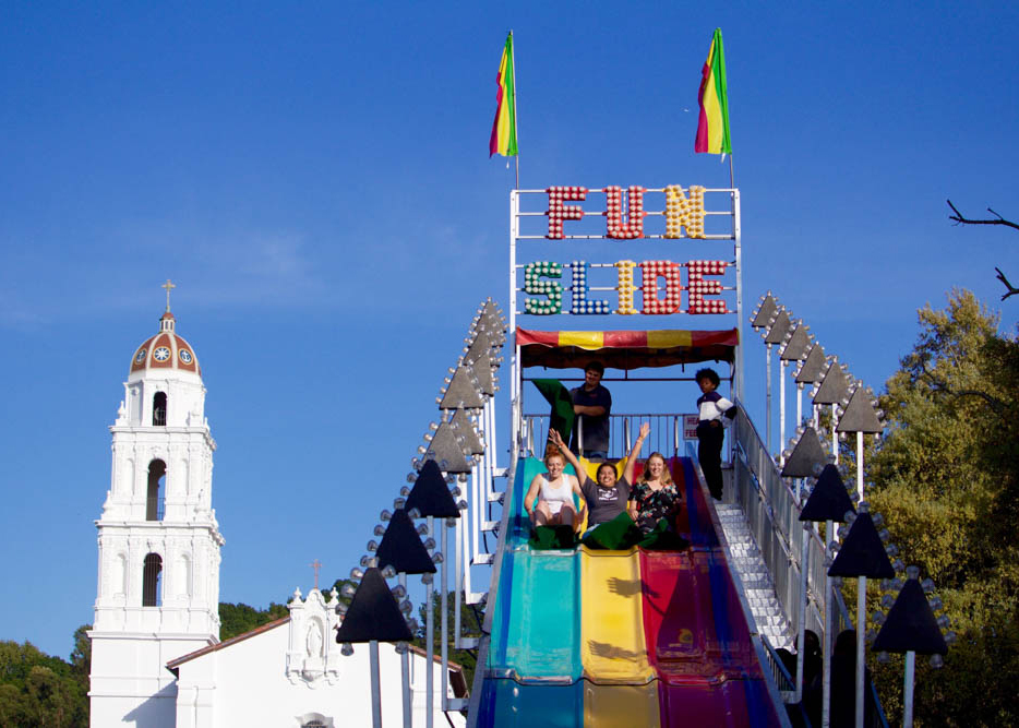 Students with their arms up sliding down a large slide during a carnival on chapel lawn