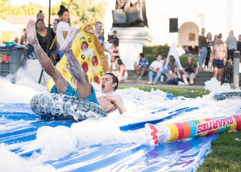 A student leaning back yelling on an inflated pool toy sliding down a foam slip and slide