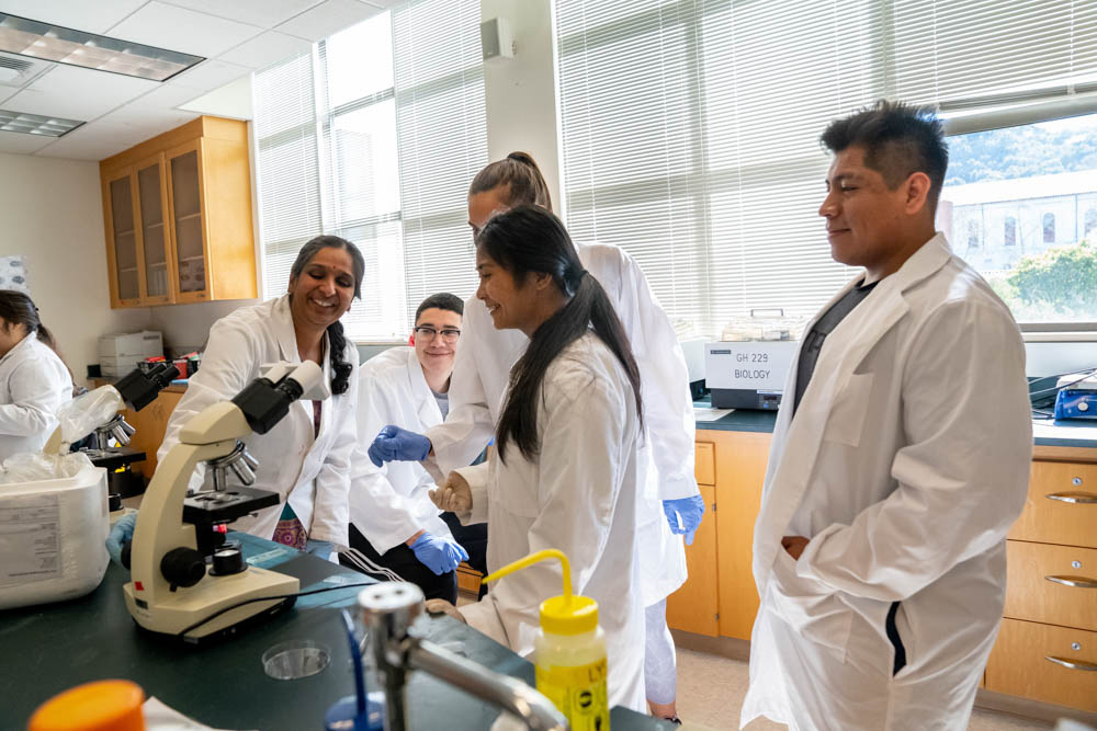 Student's in lab coats standing around a professor adjusting a microscope