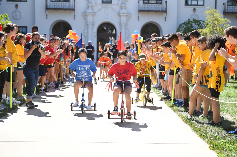 Students on tricycles racing in the first year olympics with other students cheering on the side lines