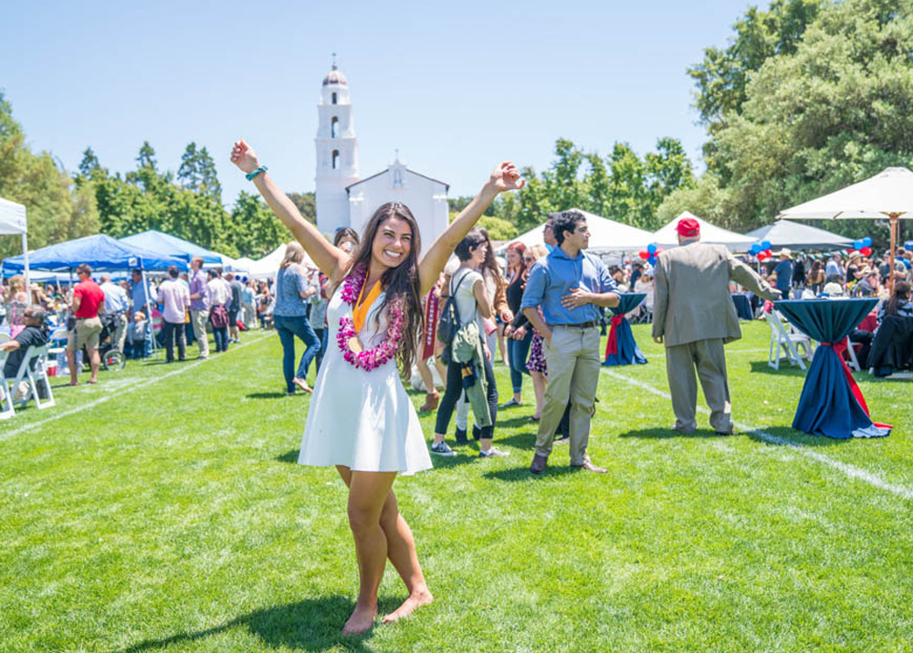 A girl with a lay on holding her arms up and smiling on chapel lawn with graduation plots behind her
