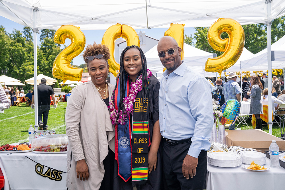 A female graduate smiling at the camera with her parents on either side and a 2019 inflatable balloon sign behind her