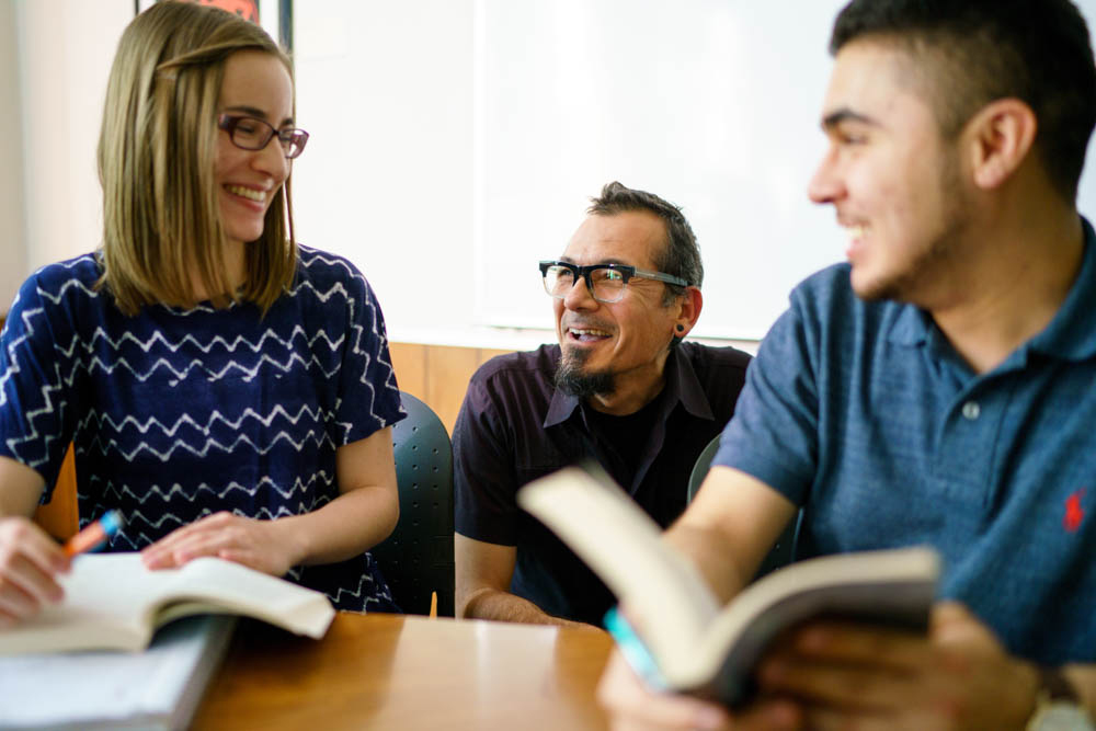 A teacher bent down between two students with books in their hand who are smiling