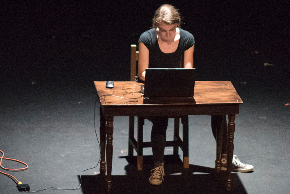 An actress performing her own play while sitting at a desk with a computer during a performance on stage of LeFevre Theatre