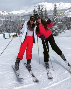 Two Girls leaning their heads together and yelling while on skis with snow covered mountains behind them