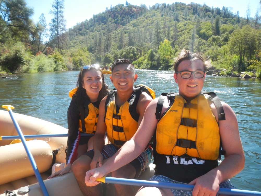 Three students with life jackets on, smiling at the camera, while sitting on a raft in a river