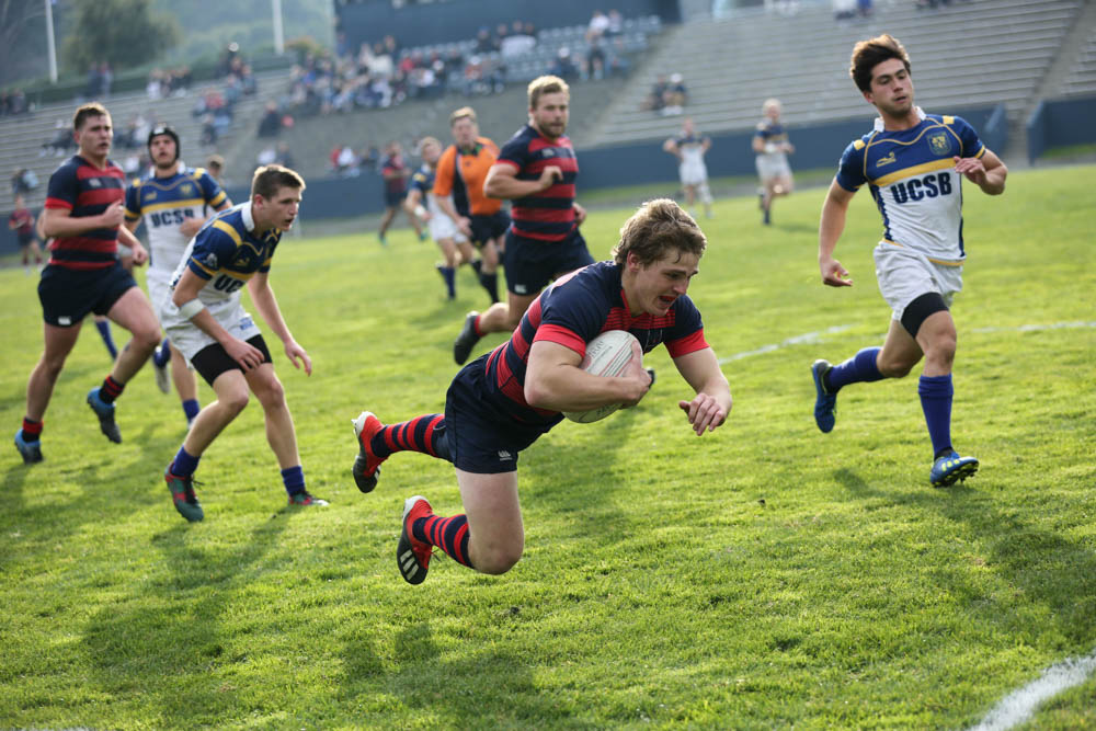 A boy diving forward with a rugby ball under his arm and guys running behind him