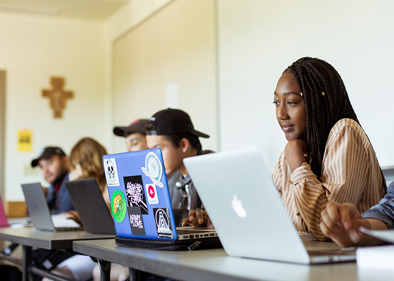 A student looking at her computer while sitting at a table with other students who are also looking at computers