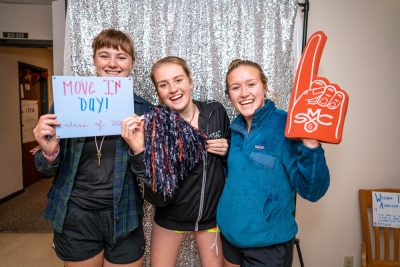 Three girls smiling in front of a glitter background. One is holding a Move In Day sign, one has a pom pom, and one has a big finger with the Gael's logo on in