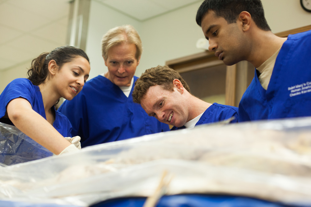 Shows students and a professor looking at a cadaver under a tarp