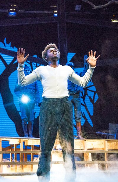 An actor on stage with his hands up and smoke below him