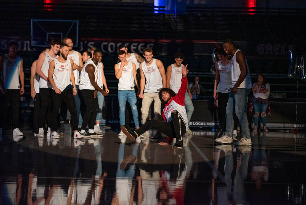 A basketball player breakdancing with the rest of the team behind him in the UCU Pavilion with the lights turned down