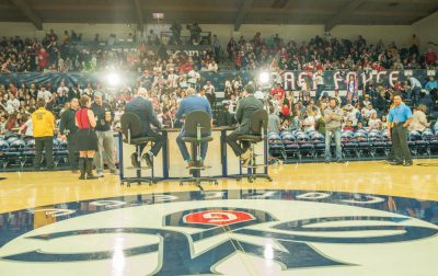 Three announcers sitting on stools of the floor of UCU Pavilion with crowds in the stands