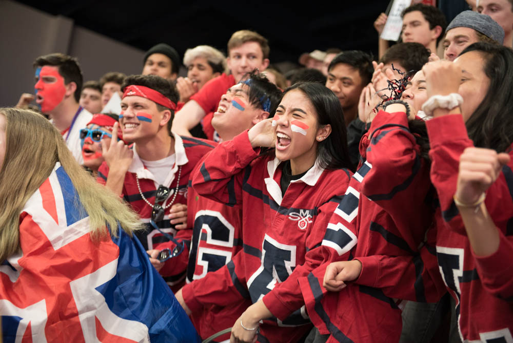 Students with painted stripes under their eyes and a letter of the word Gaels on their shirts cheering in the stands at UCU Pavilion