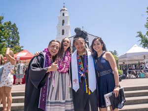 Four students with graduation gowns smiling in front of the chapel lawn